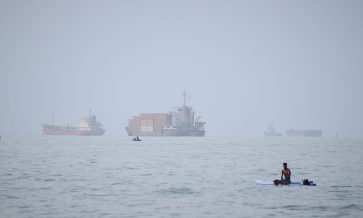 A man sits on a surfboard as a container ship and other commercial vessels appear anchored in the Strait of Hormuz off Bandar Abbas, Iran, July 27, 2026. Photo: VCG